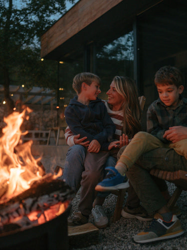 A family of four sits together by a fire pit at dusk in front of a modern house with large windows. The father and son are seated in wooden chairs, the mother sits beside them, and a young child is held by the father.