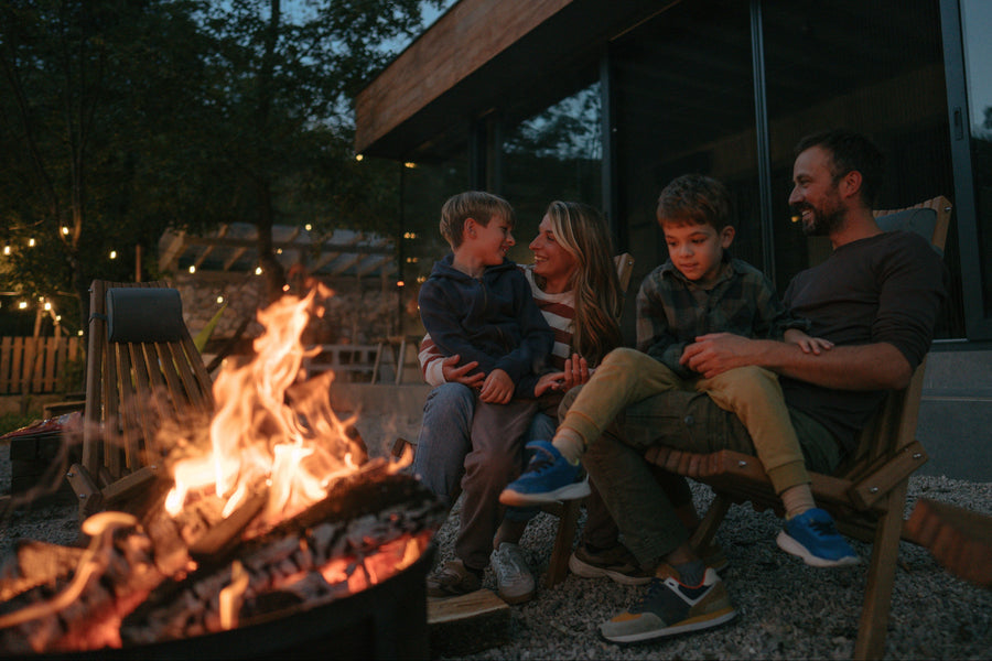 A family of four sits together by a fire pit at dusk in front of a modern house with large windows. The father and son are seated in wooden chairs, the mother sits beside them, and a young child is held by the father.